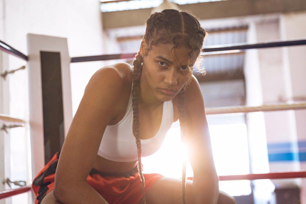 Close-up of female boxer looking at camera while relaxing in boxing ring at fitness center. Strong female fighter in boxing gym training hard.