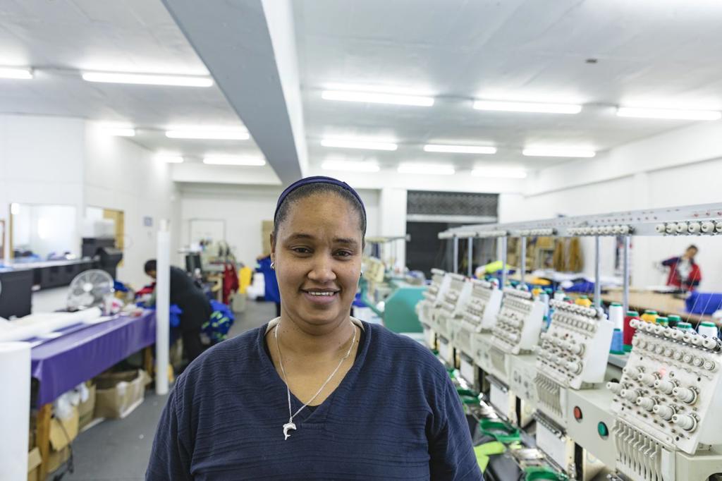 Portrait close up of a young mixed race woman standing beside rows of machines in a brightly lit sports clothing factory, looking to camera and smiling. In the background some of coworkers can be seen. They are working in a clothing factory.