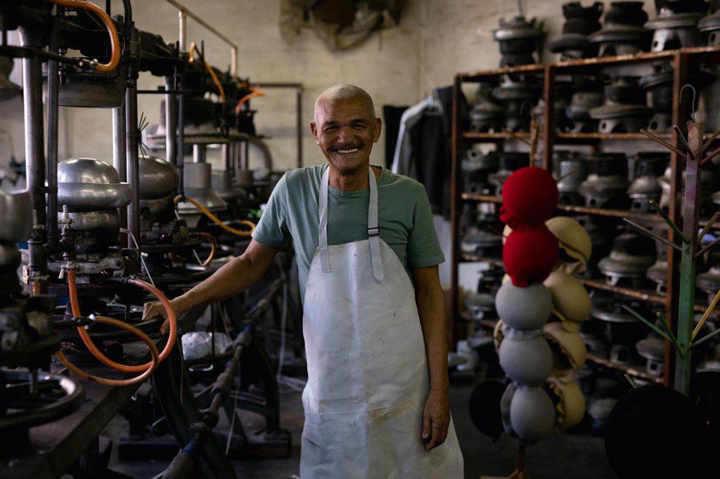 Portrait of a smiling senior mixed race man standing surrounded by traditional equipment and machinery in the workshop at a hat factory