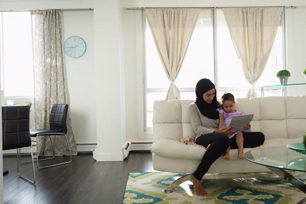 Front view of mixed race mother wearing hijab and daughter using digital tablet in living room at home 