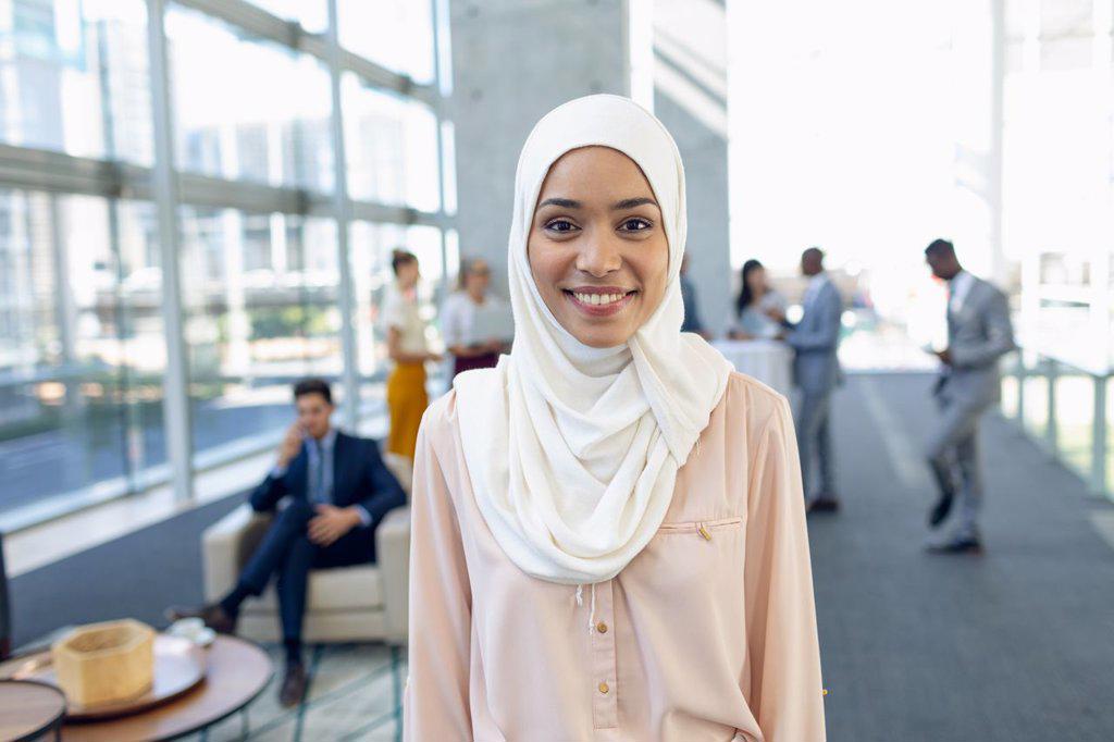 Front view of young pretty businesswoman in hijab looking at camera while standing in modern office. Behind her, colleagues interacting with each other.