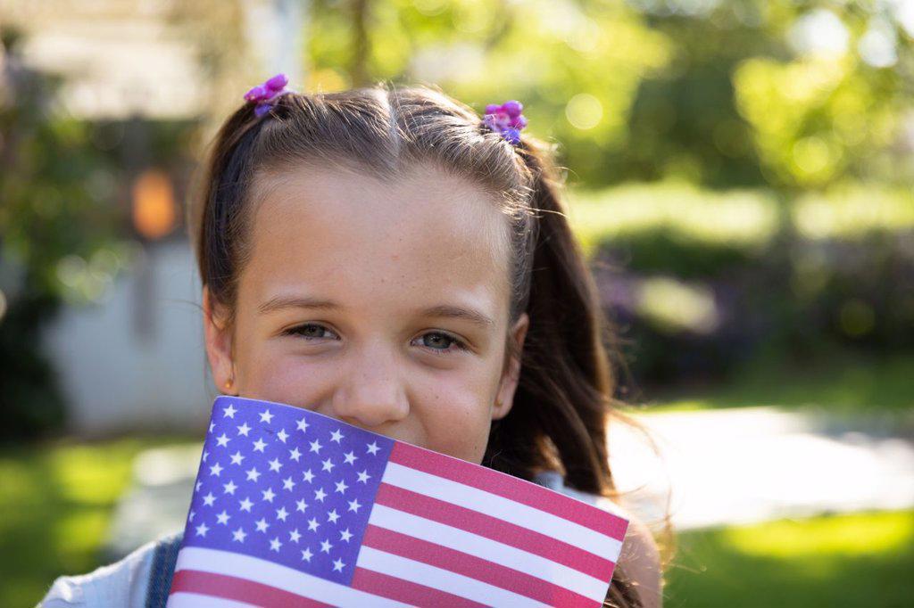 Portrait close up of a young Caucasian girl with long brown hair and blue eyes outside in a sunny garden holding a US flag and smiling to camera. Family enjoying time at home, lifestyle concept