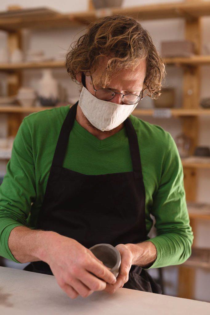 Portrait of caucasian man wearing face mask at pottery studio. small creative business during covid 19 coronavirus pandemic.