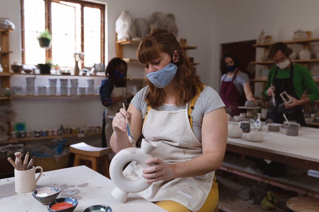 Caucasian female potter in face mask working in pottery studio. wearing apron, working at a working table with her friends in the back. small creative business during covid 19 coronavirus pandemic.