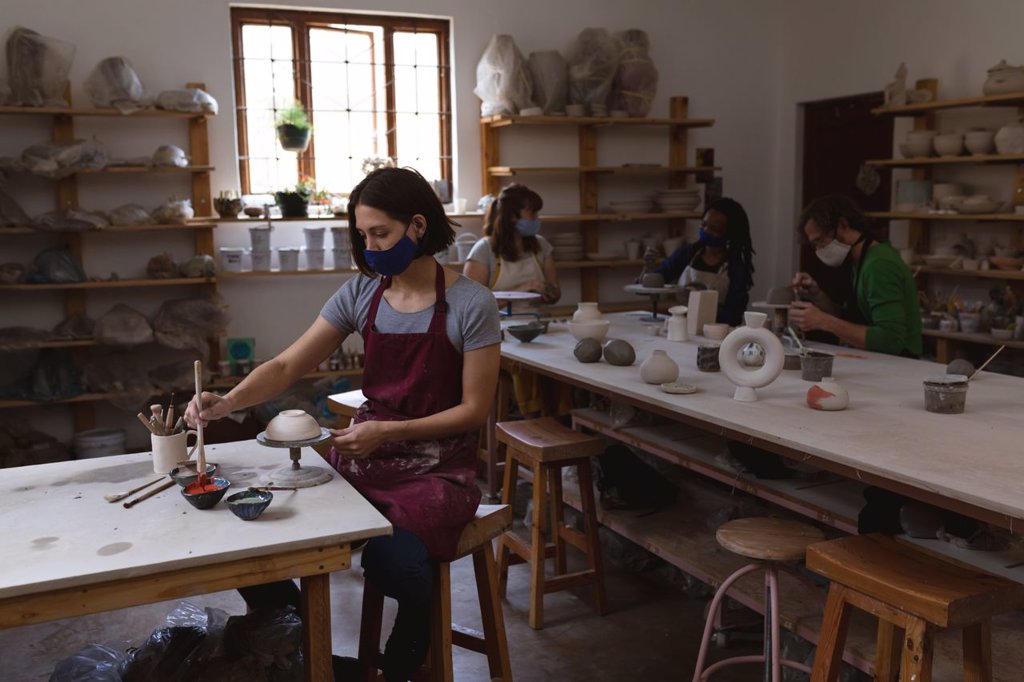 Caucasian female potter in face mask working in pottery studio. wearing apron, working at a working table with her friends in the back. small creative business during covid 19 coronavirus pandemic.