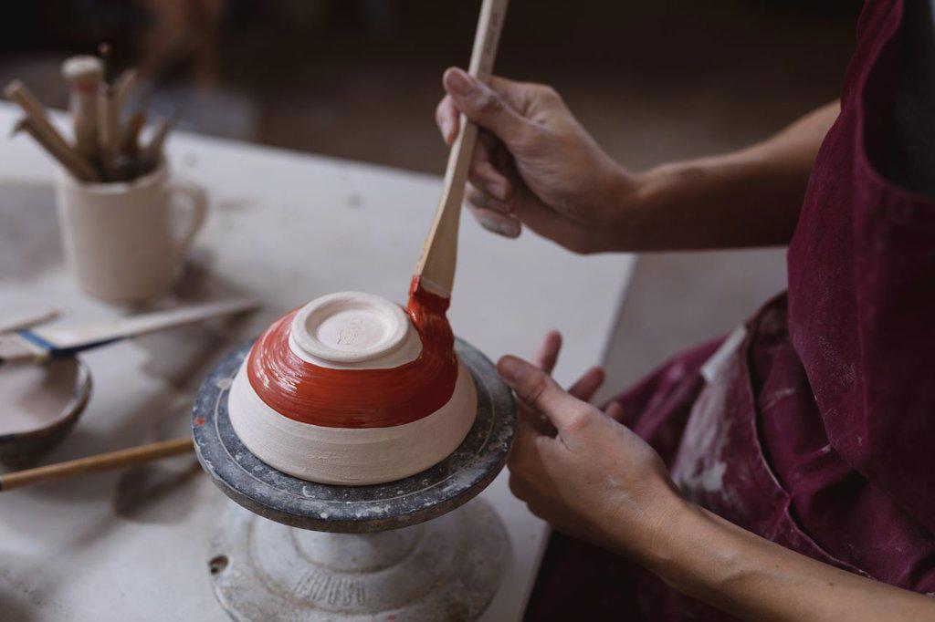 Caucasian female potter in face mask working in pottery studio. wearing apron, working at a potters wheel, painting a bowl. small creative business during covid 19 coronavirus pandemic.