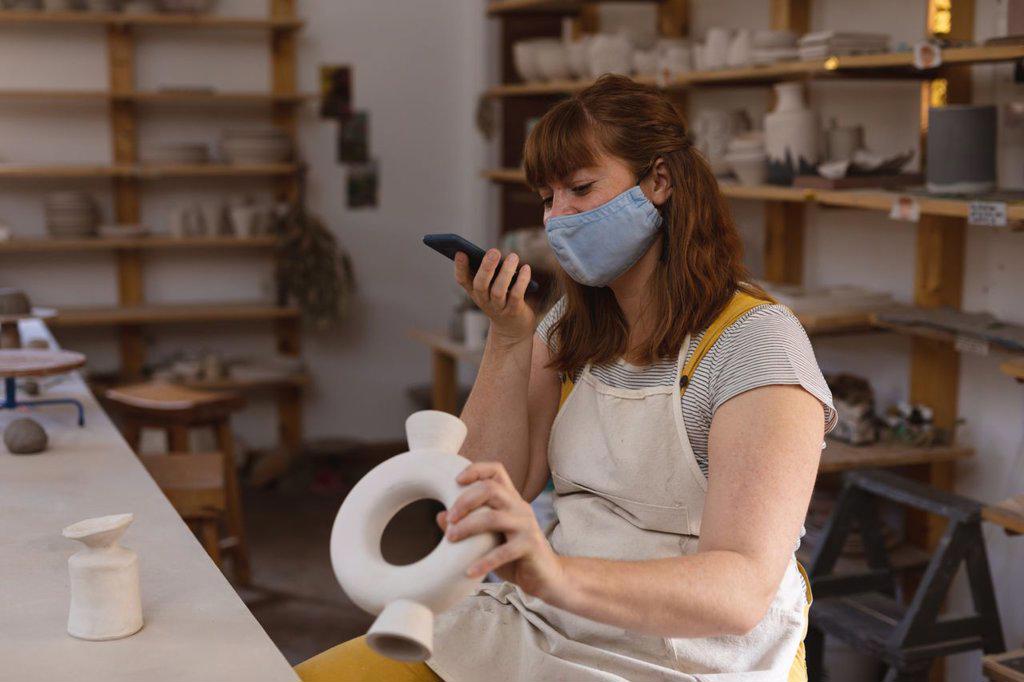 Caucasian female potter in face mask working in pottery studio. wearing apron, talking on the phone. small creative business during covid 19 coronavirus pandemic.