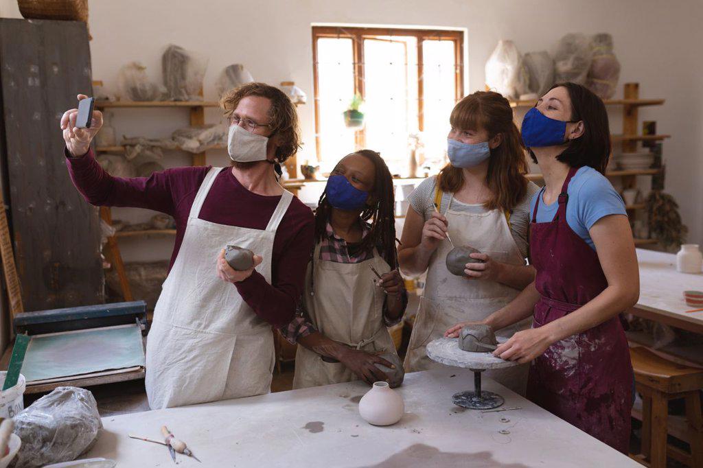 Multi-ethnic group of potters in face masks working in pottery studio. wearing aprons, painting plates, taking a selfie together. small creative business during covid 19 coronavirus pandemic.