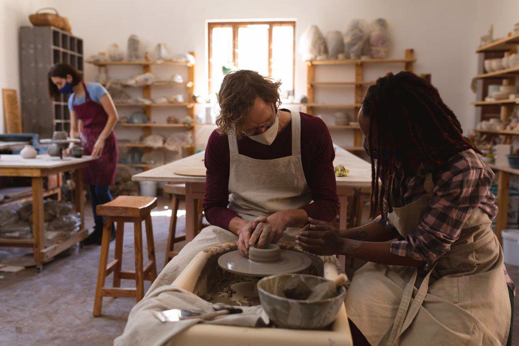 Caucasian male and mixed race female potters in face masks working in pottery studio. wearing aprons, working at a potters wheel. small creative business during covid 19 coronavirus pandemic.