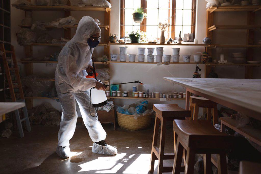 Caucasian male cleaner in protective clothes working in pottery studio. disinfecting the whole place. small creative business during covid 19 coronavirus pandemic.