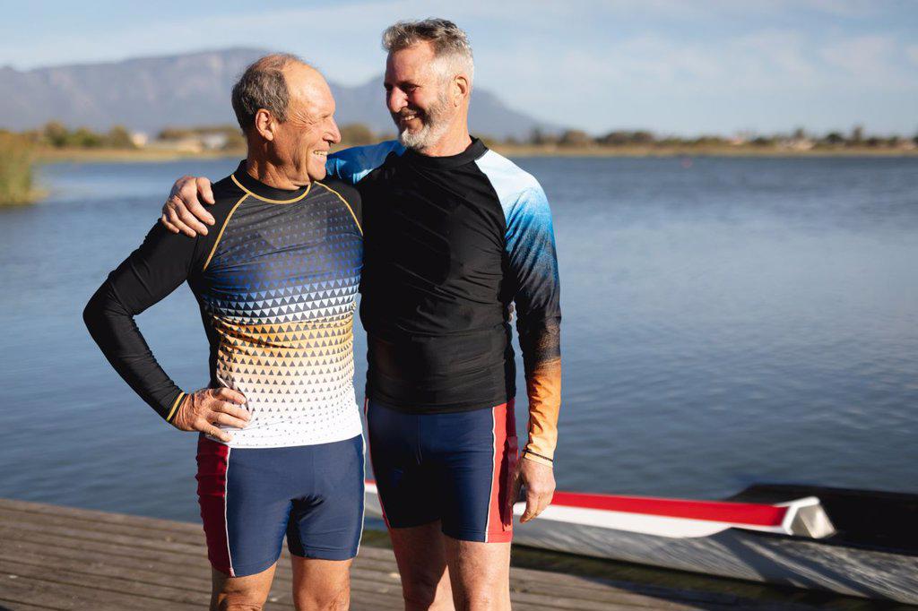 Two senior caucasian male rowers smiling looking at each other while standing on the wooden dock. retirement sports and active senior lifestyle.