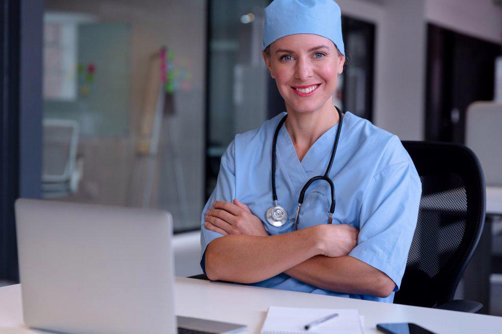 Portrait of smiling caucasian female doctor sitting at desk wearing scrubs using laptop computer. medical professional at work.