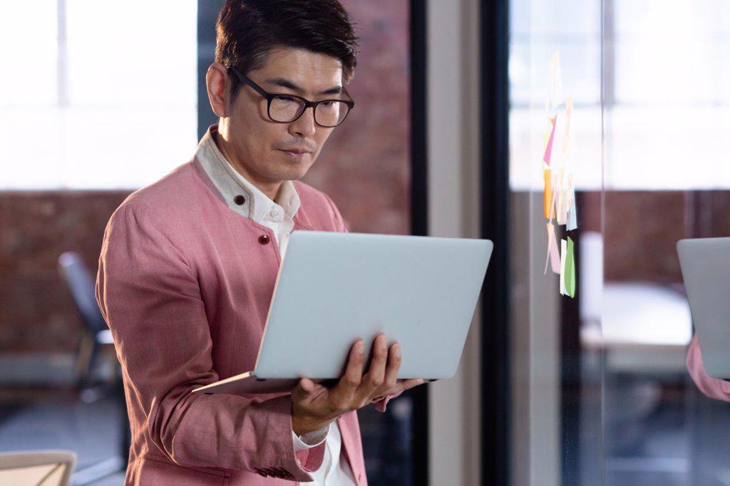 Stylish asian businessman standing and using laptop computer. business person at work in modern office.