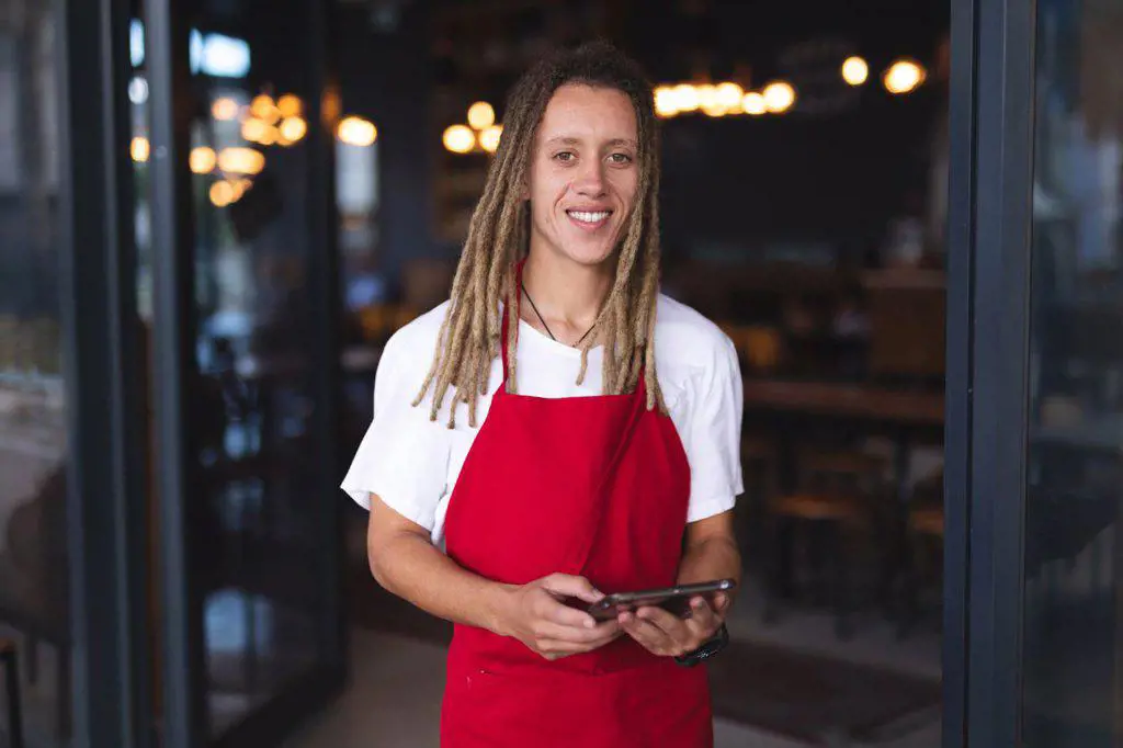Portrait of happy mixed race male barista with dreadlocks standing in doorway of cafe holding tablet. independent small business in a city.