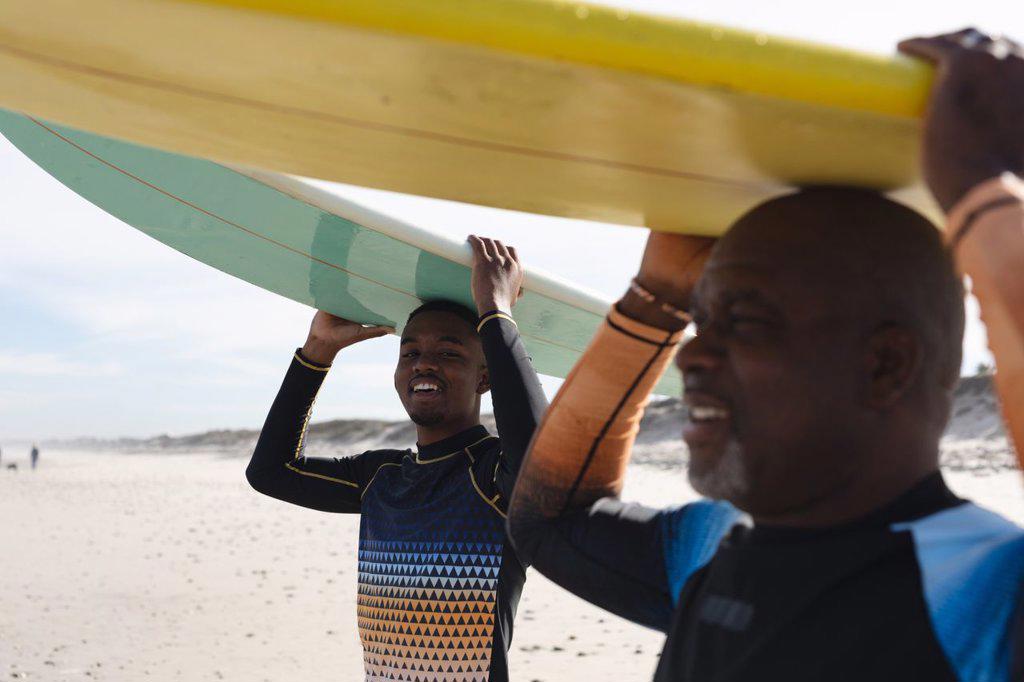 African american father and son carrying surfboards on their heads at the beach. summer beach holiday and leisure concept.