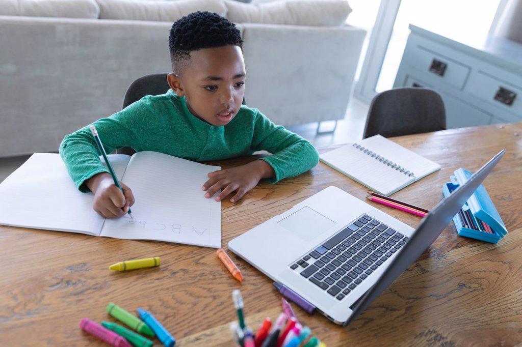 African american boy in online school class, using laptop and writing in his notebook. at home in isolation during quarantine lockdown.