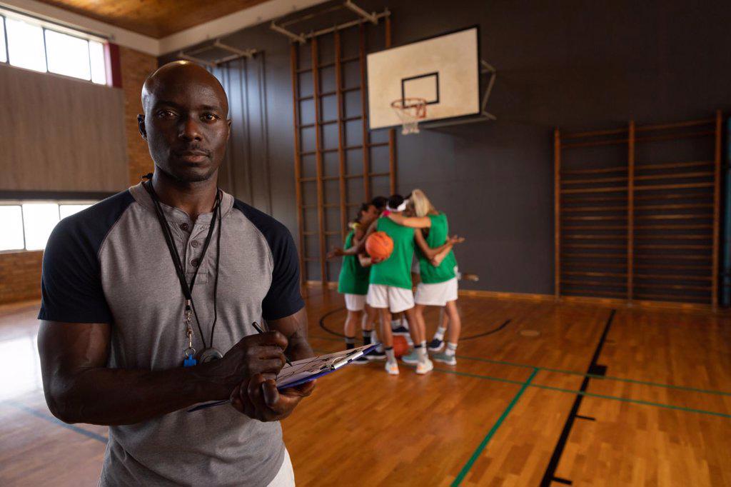 Portrait of african american male basketball coach holding clipboard with team in background. basketball, sports training at an indoor court.