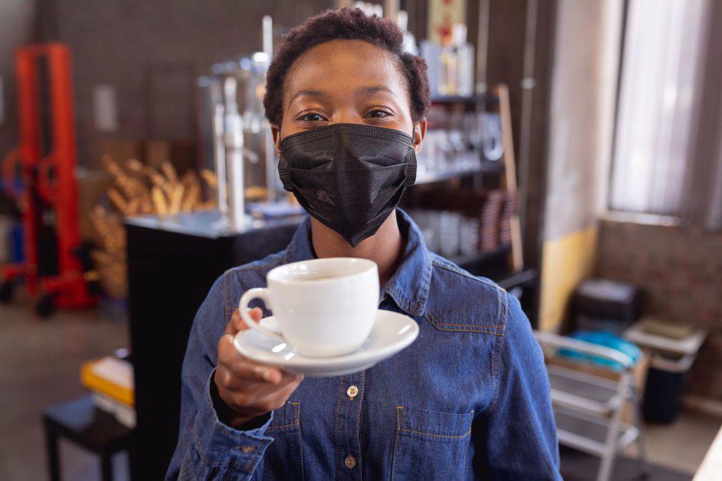 Portrait of african american woman wearing face mask holding a coffee cup at a cafe. health protection and safety during covid-19 pandemic concept