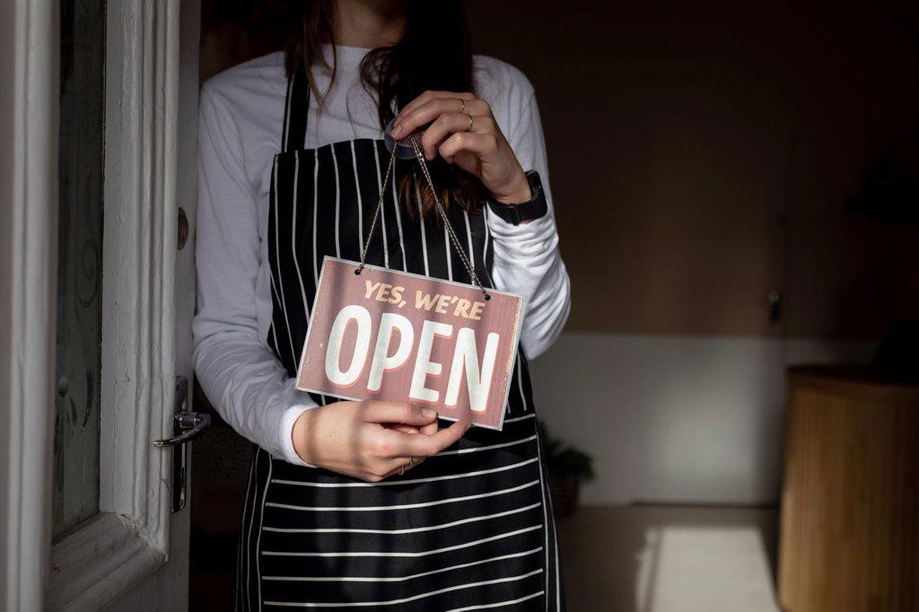 Midsection of caucasian waitress wearing striped apron, standing in doorway, holding open sign. small independent cafe business.