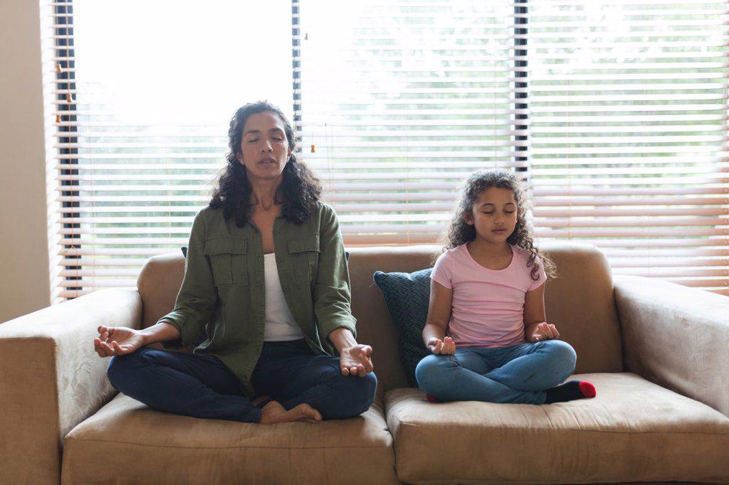 Mixed race mother and daughter sitting on sofa and meditating. domestic lifestyle and spending quality time at home.