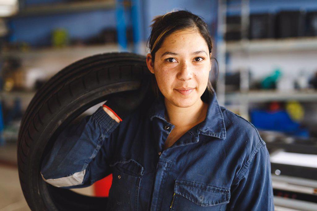 Mixed race female car mechanic wearing overalls, holding tire, looking at camera. independent business owner at car servicing garage.