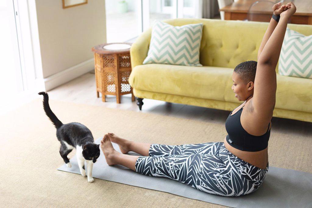Happy african american plus size woman practicing yoga on mat at home with cat. fitness and healthy, active lifestyle.