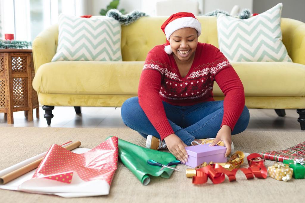 Happy african american plus size woman in santa hat wrapping presents at home. christmas, festivity and tradition concept.