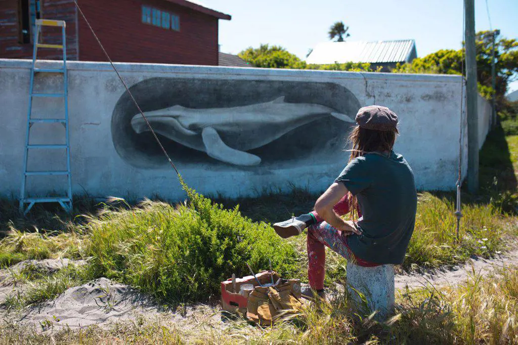Male artist sitting while admiring his beautiful whale mural on wall during sunny day. street art and skill.