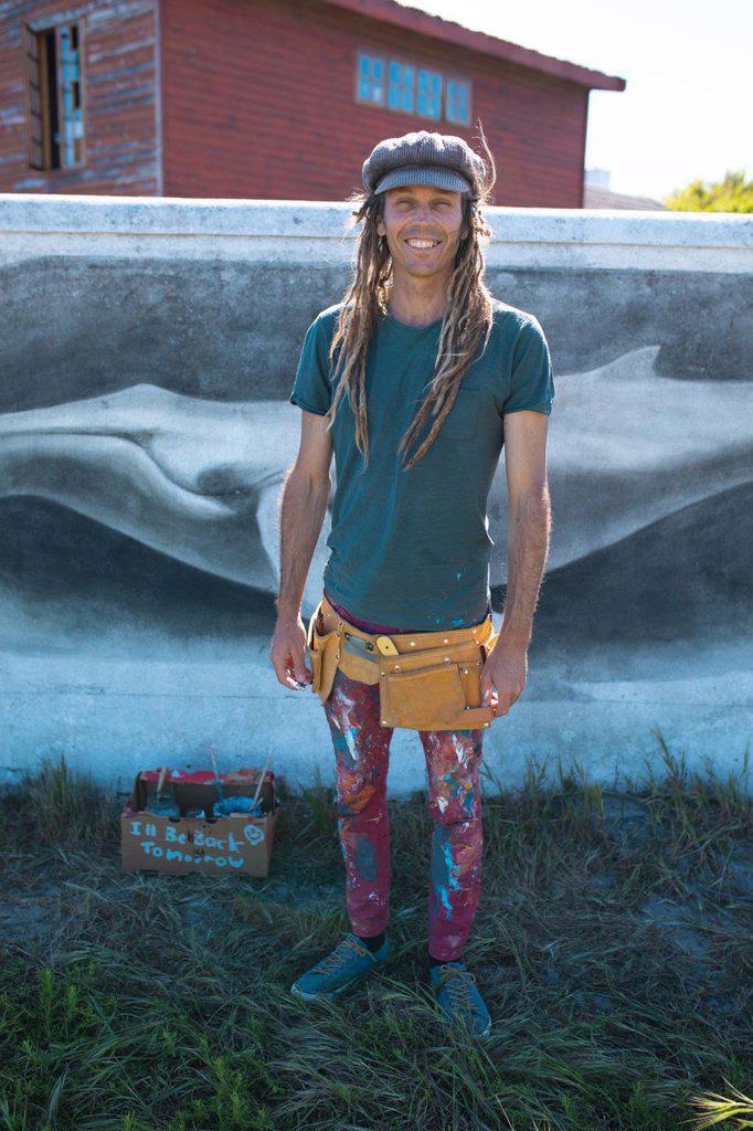 Smiling portrait of male hipster artist with messy clothing standing against whale mural painting. street art and skill.