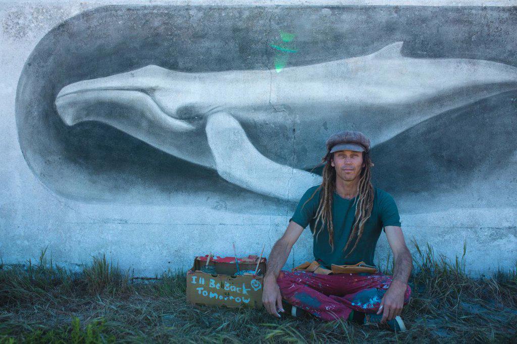 Proud male hipster artist sitting cross-legged in front of his whale mural painting on wall. street art and skill.