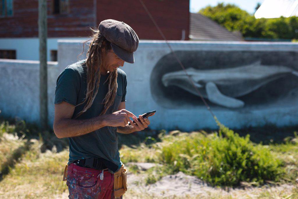 Male mural artist using smart phone while standing near whale painting on wall during sunny day. technology, street art and skill.