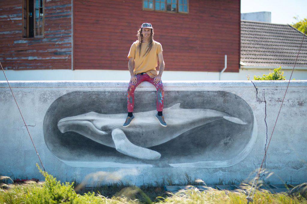 Portrait of male artist sitting on wall with whale mural painting against house. street art and skill.