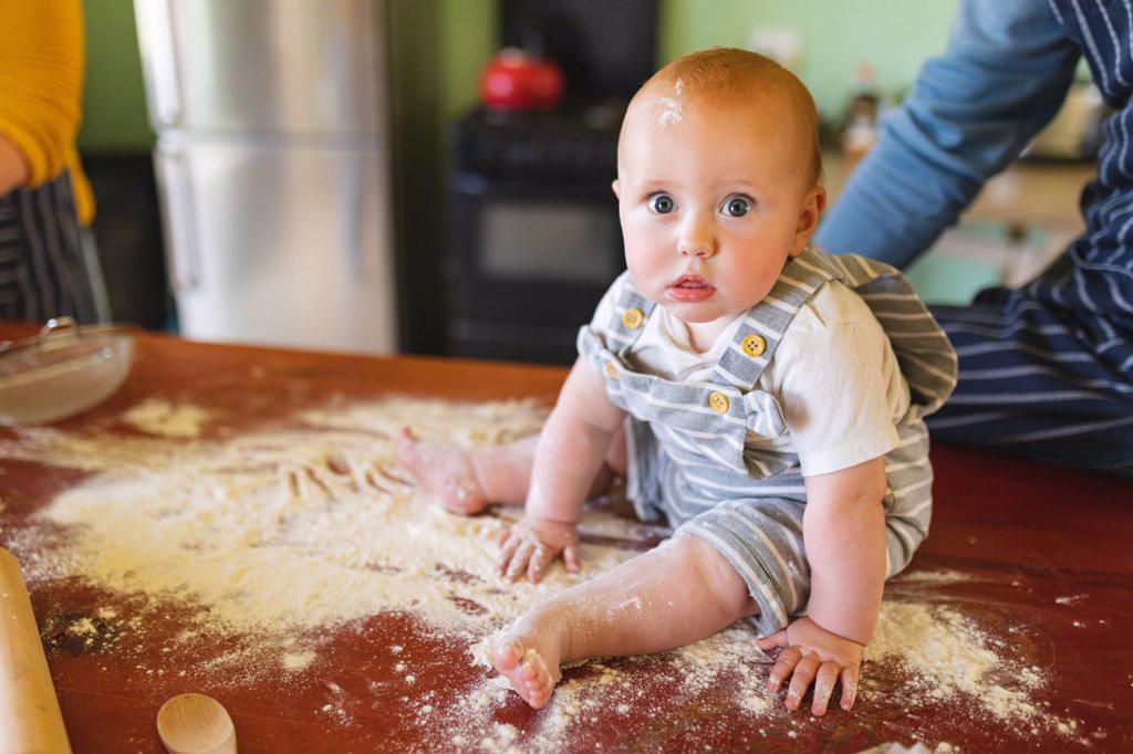 Portrait of cute baby playing while sitting on flour over table with parents in kitchen. innocence and family.