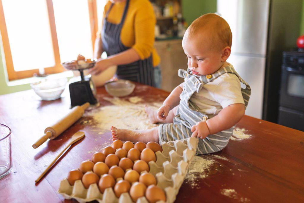 Cute baby playing with egg carton on wooden table while mother preparing food in kitchen. innocence, family and healthy eating.