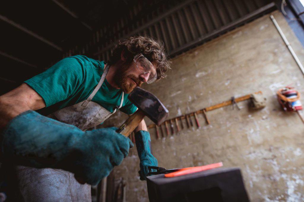 Bearded caucasian blacksmith in protective workwear forging with hammer on anvil in industry. forging, metalwork and manufacturing industry.