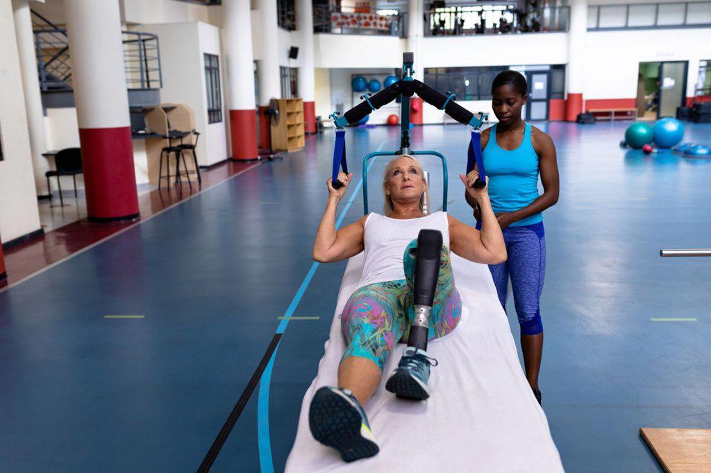 Front view of African-american female trainer assisting disabled Caucasian active senior woman to exercise in sports center. Sports Rehab Centre with physiotherapists and patients working together towards healing