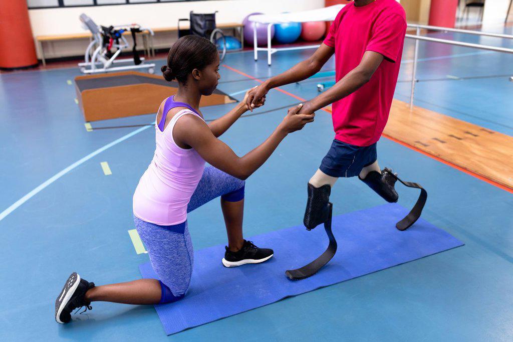 Side view of African-american physiotherapist helping disabled African-american man walk with prosthetic leg in sports center. Sports Rehab Centre with physiotherapists and patients working together towards healing