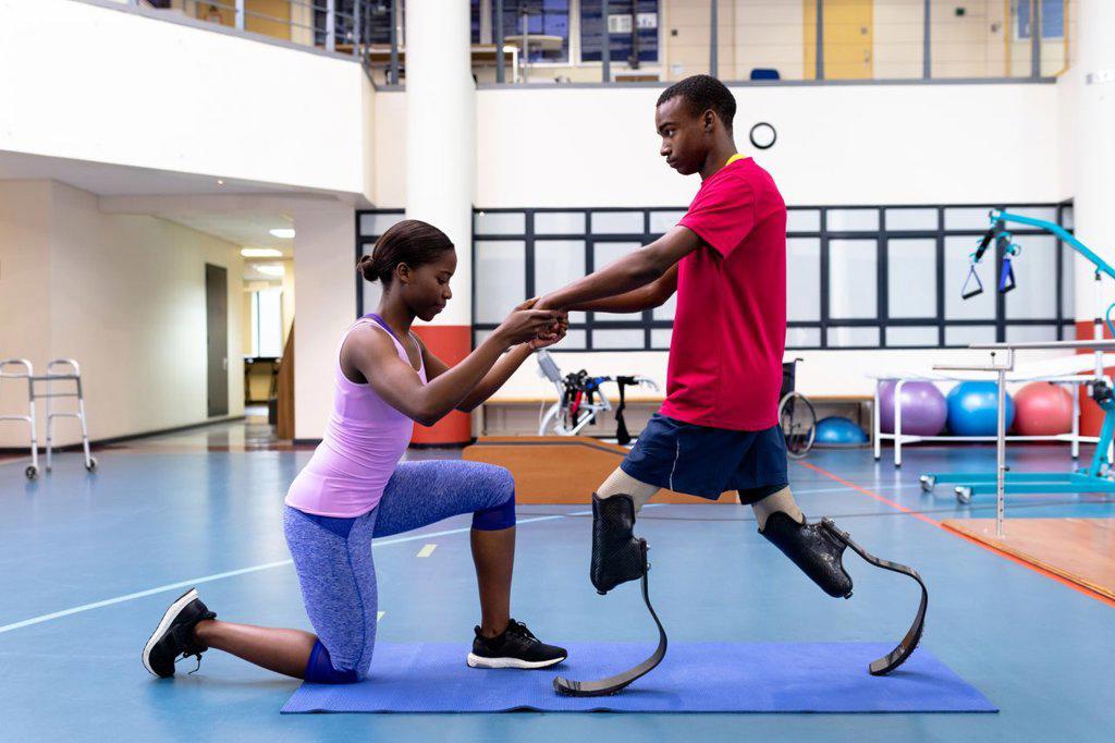 Side view of African-american physiotherapist helping disabled African-american man walk with prosthetic leg in sports center. Sports Rehab Centre with physiotherapists and patients working together towards healing
