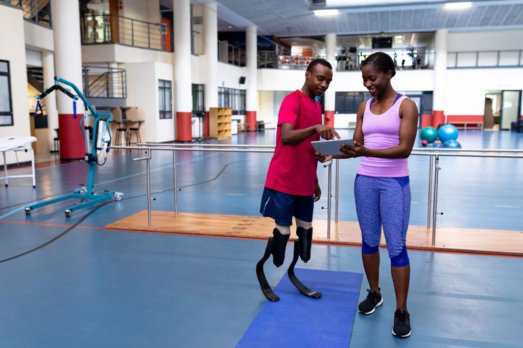 Front view of African-american female trainer and disabled African-american man discussing over digital tablet in sports center. Sports Rehab Centre with physiotherapists and patients working together towards healing