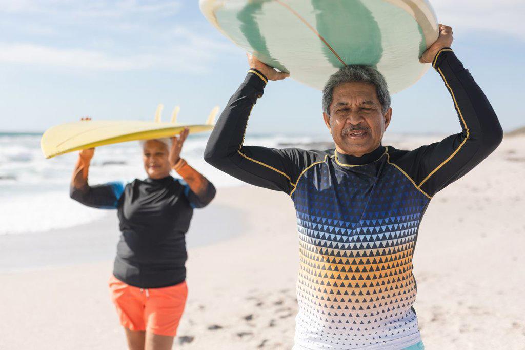 Multiracial senior couple enjoying retirement together while carrying surfboards over heads at beach. water sport and active lifestyle.