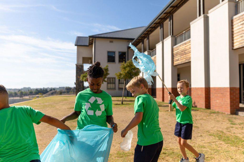 Multiracial elementary schoolboys cleaning ground by school building during sunny day. unaltered, sustainable lifestyle, education, teamwork, cleaning, responsibility and recycling concept.