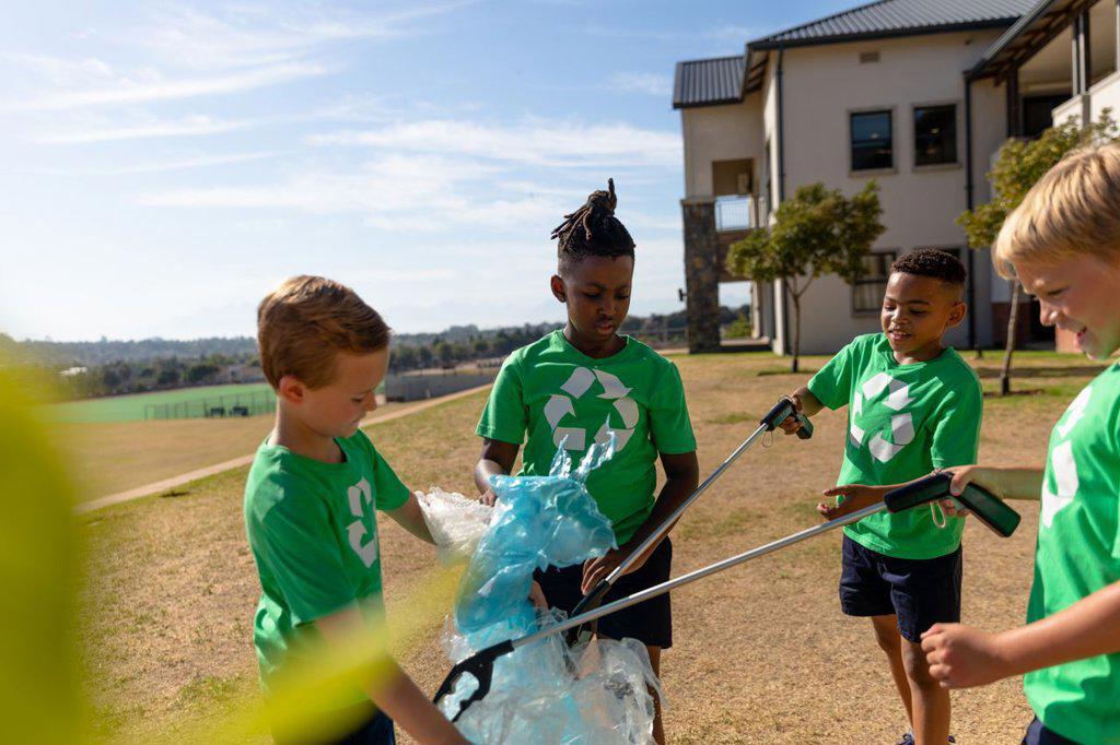Multiracial elementary schoolboys recycling plastic in garbage bag at school ground. unaltered, sustainable lifestyle, teamwork, cleaning, responsibility and recycling concept.