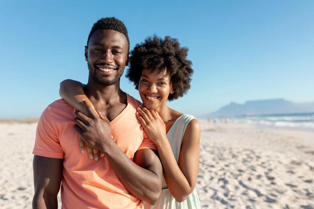 Portrait of smiling african american woman embracing boyfriend from behind at beach on sunny day. unaltered, lifestyle, love, togetherness and holiday concept.