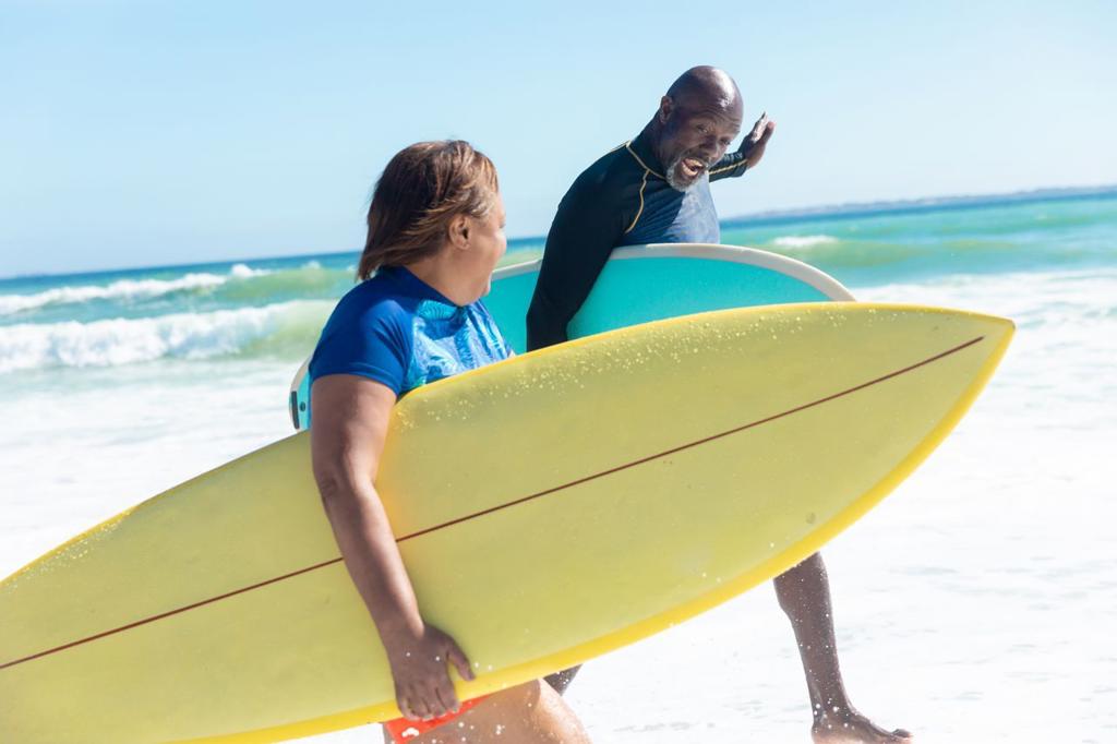 African american retired senior couple talking while carrying surfboards at beach on sunny day. unaltered, togetherness, active lifestyle, aquatic sport and holiday concept.