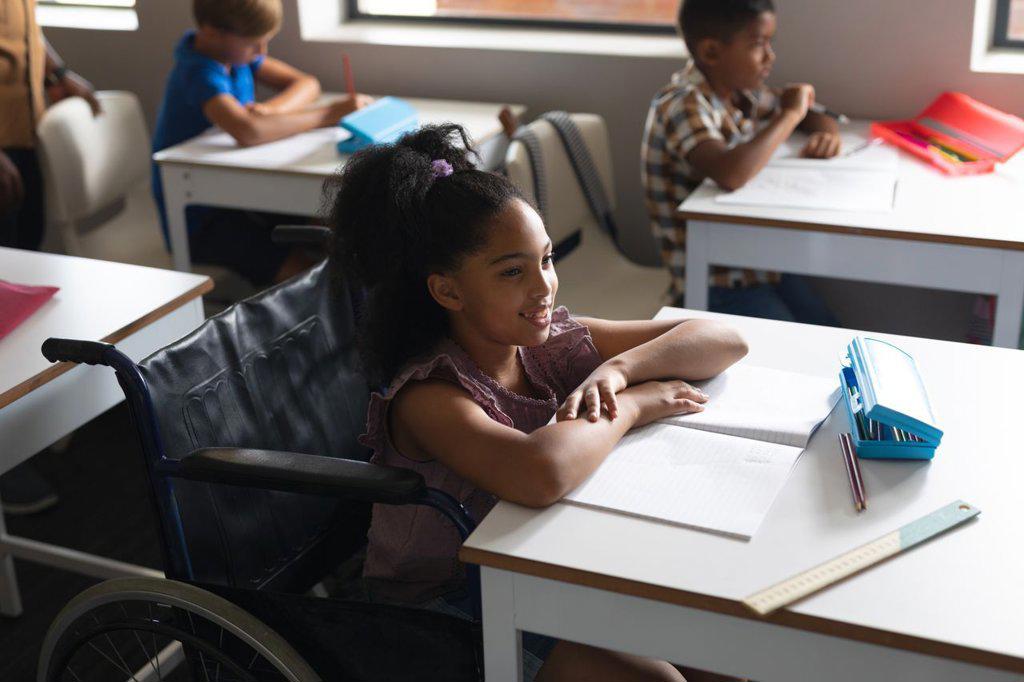 Smiling biracial elementary schoolgirl sitting on wheelchair at desk in classroom. unaltered, education, childhood, learning, disability, physical disability and school concept.