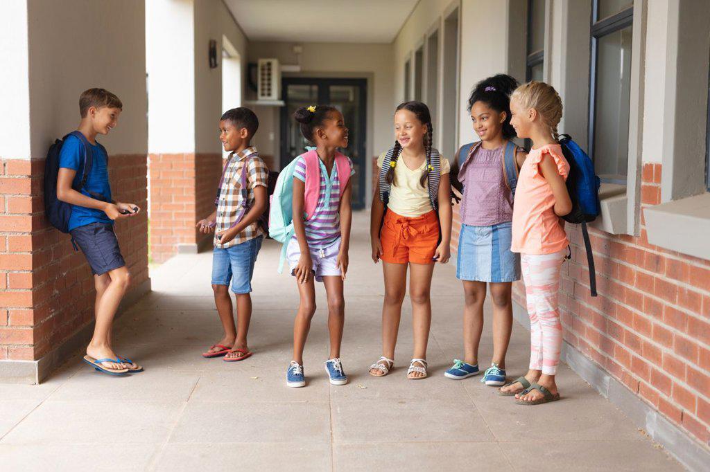 Full length of multiracial elementary school students talking while standing in school corridor. unaltered, education, childhood, together and school concept.