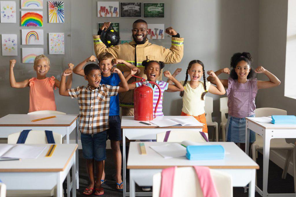 Multiracial elementary students and african american young male teacher flexing muscles. unaltered, education, firefighter, safety, protection, teaching and school concept.