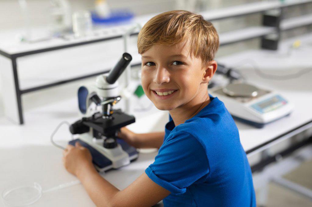 Portrait of smiling caucasian elementary schoolboy with microscope sitting at desk in laboratory. unaltered, education, childhood, learning, science, stem and school concept.