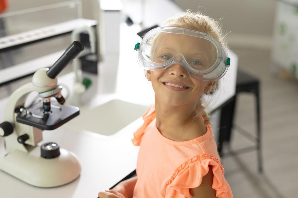Portrait of smiling caucasian elementary schoolgirl with protective glasses sitting in laboratory. unaltered, education, childhood, learning, science, stem and school concept.