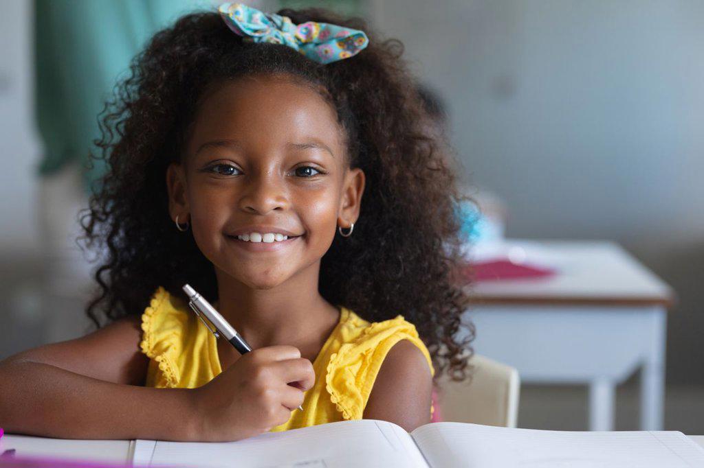 Portrait of smiling african american elementary schoolgirl with curly hair sitting at desk in class. unaltered, education, childhood, learning, studying, book and school concept.
